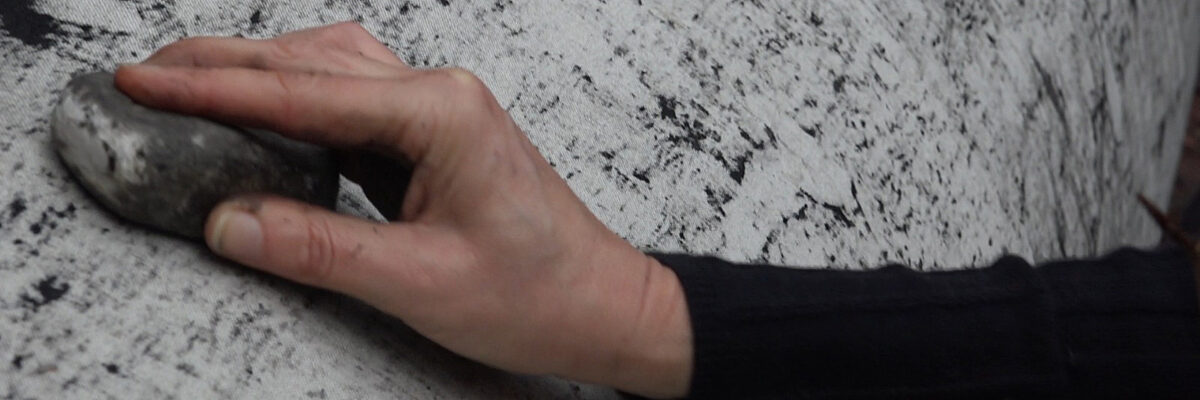 Een hand wrijft met een steen op de schors van een boom die omwikkeld is met witte stof, waardoor tekeningen ontstaan op de stof. 

A hand rubs a stone on the bark of a tree that is covered in white cloth, creating marks on the cloth.
