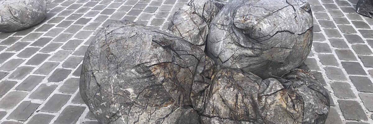 Steenachtige vormen ter grootte van een mens liggen op straat, op kasseien naast het water. 

Rock-like shapes the size of a person lie on the street, on a cobbled are next to a bassin of water.