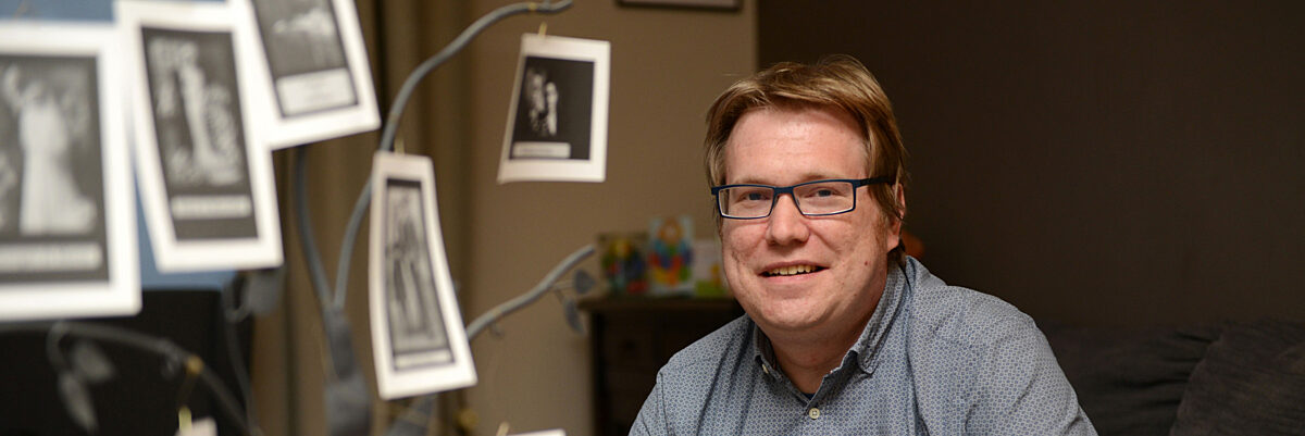 Man met blauw hemd zit op een grijze zetel en lacht naar de camera, tussen hem en de camera hangen (onscherpe) familiefoto's. 

Man wearing a blue shirt sitting on a grey sofa, smiling into the camera, with blurry family pictures filling the other half of the frame.