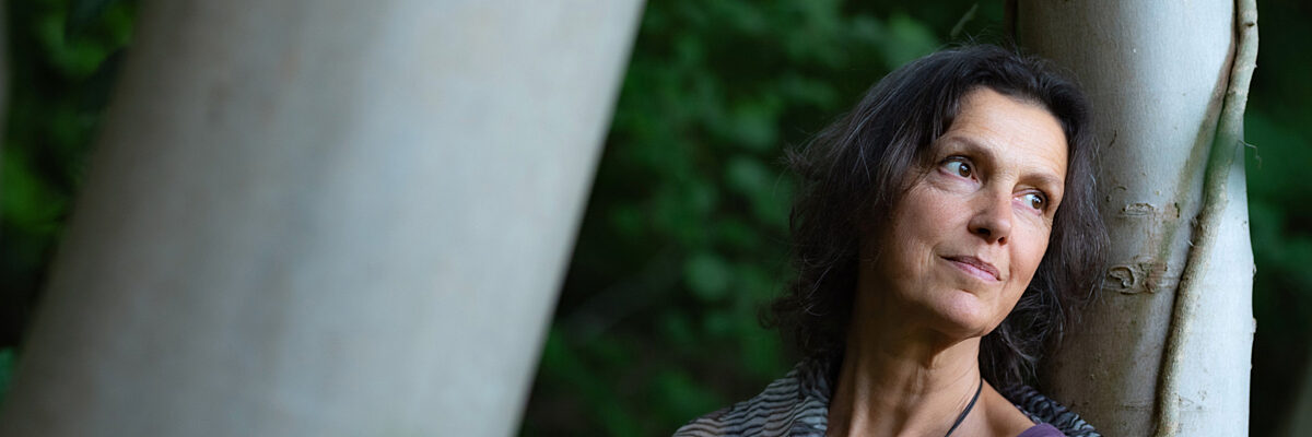 Vrouw met een paars topje staat in een bos, haar rug leunend tegen een witte boom, en kijkt dromerig naar links. 

Woman wearing a purple top stands in a forest, leaning her back on a white tree, looking dreamily to her left.