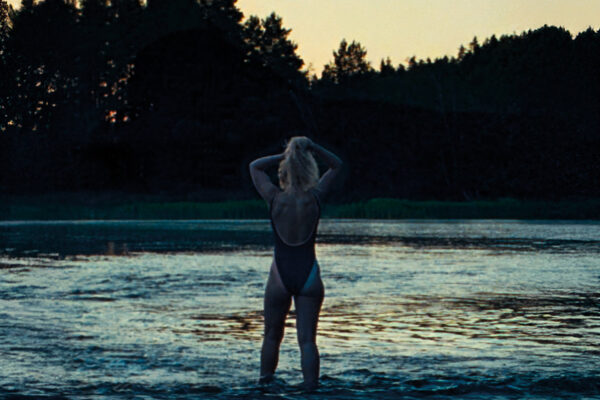 Blonde vrouw met rug naar camera staat met badpak aan met haar voeten in een meer in de bossen.

Blonde woman with her back to the camera, wearing a bathing suit, standing with her feet in a forest lake.