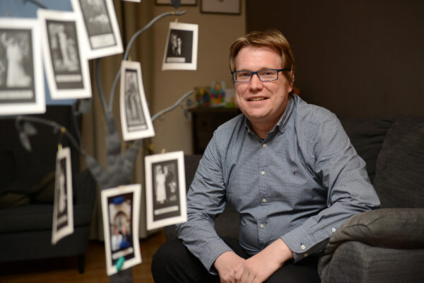 Man met blauw hemd zit op een grijze zetel en lacht naar de camera, tussen hem en de camera hangen (onscherpe) familiefoto's. 

Man wearing a blue shirt sitting on a grey sofa, smiling into the camera, with blurry family pictures filling the other half of the frame.