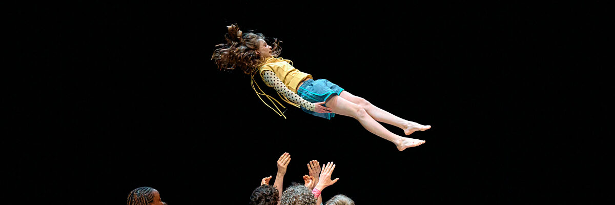 Een danser springt in de lucht terwijl een groep mensen beneden omhoog reikt op een donker, schemerig verlicht podium.

A dancer leaps mid-air while a group of people below reaches up toward them on a dark, dimly lit stage.