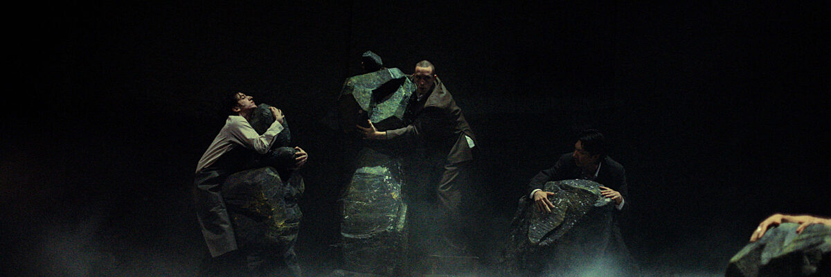 Vier performers in donkere kostuums houden grote, getextureerde stenen vast op een donker, rokerig podium, een sfeervolle theaterscène.

Four actors in dark costumes hold large, textured rocks on a dimly lit, smoky stage, creating an intense dramatic atmosphere.