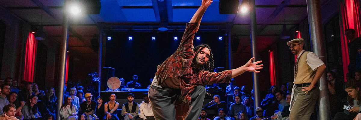 Een danser in een patronenhemd treedt op op een houten dansvloer met een aandachtig publiek dat op de achtergrond zit.

A dancer in a patterned shirt performs on a wooden stage with an attentive audience seated in the background.
