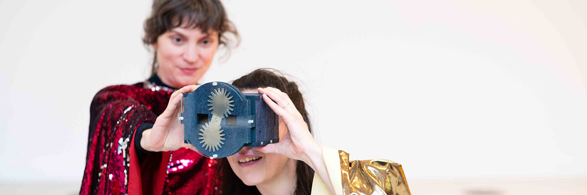 Twee personen, één in een rode paillettenjas en een andere in een gouden cape, houden een apparaat voor hun gezicht in een lichte studio.

Two people, one in a red sequin jacket and another in a gold cape, hold a device to their face in a bright studio.