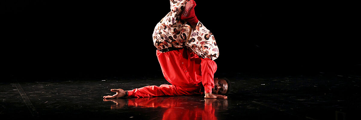 Een danser in een rood patronenpak voert een kopstand uit op een donker podium met dramatische verlichting en reflecties.

A dancer in a red patterned outfit performs a headstand on a dark stage with dramatic lighting and reflections.