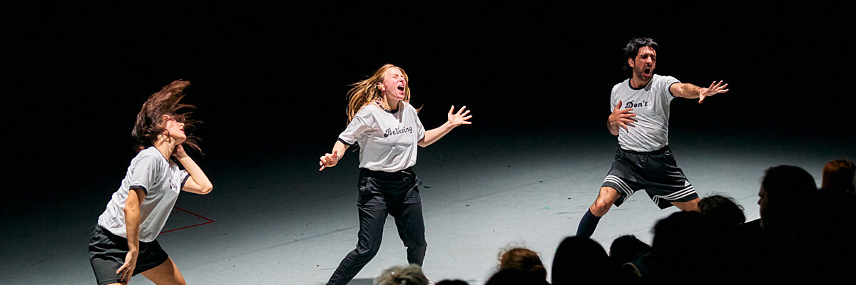 Drie dansers treden op op een donker podium met publiek op de achtergrond, twee in witte shirts en één in zwarte short.

Three dancers perform on a dark stage with an audience in the background, two in white shirts and one in black shorts.