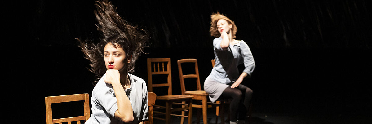Twee vrouwen op een donker podium, één zittend op een stoel met kin in hand, de andere zit verder achterin op een stoel met wapperend haar.

Two women on a dark stage, one seated on a chair with chin in hand, the other sitting further on a chair with hair blowing.