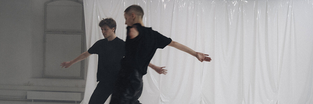 Twee mannen in zwart dansen energiek in een witte studio met plastic doek achtergrond en boogvenster.

Two men in black clothing dance energetically in a white studio with plastic sheet backdrop and arched window.