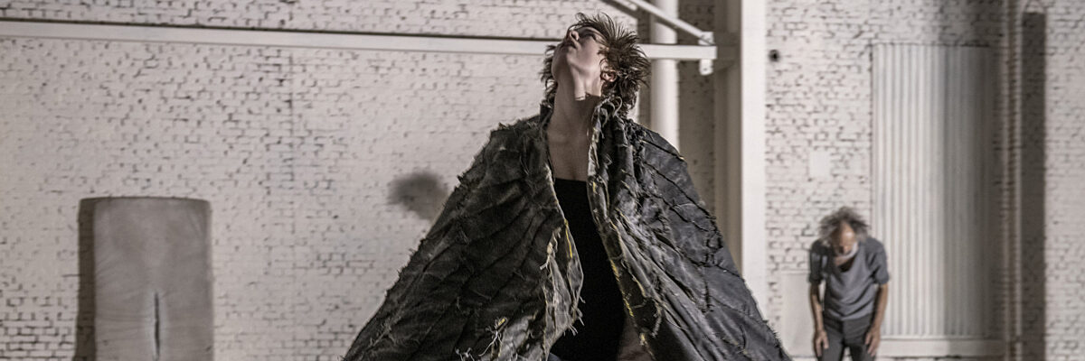 Een persoon in een versleten zwart vleugelachtig kostuum loopt in een witte bakstenen studio met een andere persoon op de achtergrond.

A person in a tattered black wing-like costume walks in a white brick-walled studio with another person in the background.