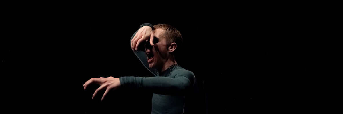 Een man in een groen shirt en donkere short hurkt op een zwarte achtergrond, één hand op gezicht, de andere uitgestrekt.

A man in a green long-sleeve shirt and dark shorts crouches on a black background, one hand on face, the other extended.