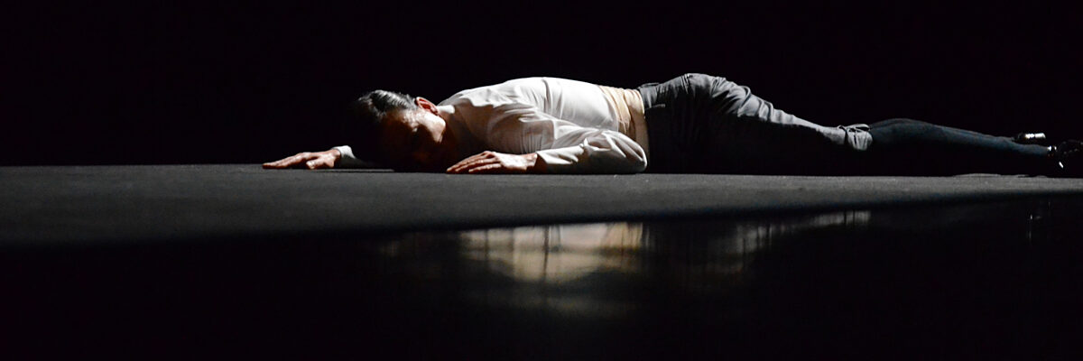 Een persoon ligt op de buik op een donker podium met uitgestrekte armen, gekleed in een wit shirt en donkere broek, onder subtiele, gerichte verlichting.

A person lies on their belly on a dark stage with arms stretched out, wearing a white shirt and dark pants, under subtle, focused lighting.