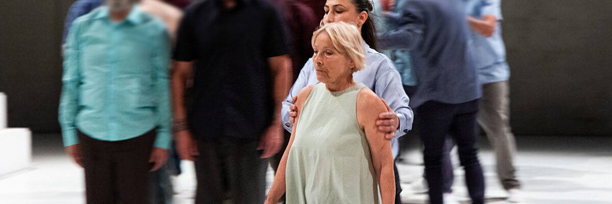 Een groep mensen in casual kleding loopt op een lichtkleurig podium, met een vrouw in een lichte groene jurk duidelijk op de voorgrond.

A group of people in casual clothing walks on a light-colored stage, with a woman in a light green dress prominently in the foreground.
