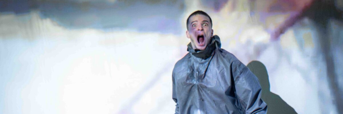 Een persoon in een glanzend metalen pak en gasmasker schreeuwt met open mond op een donker podium met kleurrijke, sfeervolle lichteffecten.

A person in a shiny metallic suit and gas mask screams with mouth wide open on a dark stage with colorful, atmospheric lighting.