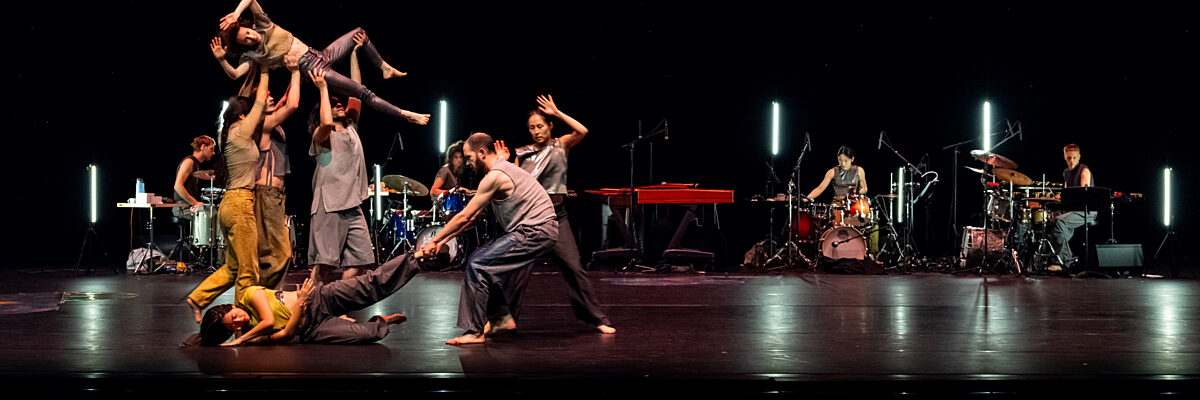 Dansers treden op op een donker podium met een live band op de achtergrond, waaronder één danser die door anderen wordt opgetild in een dynamische groepspose.

Dancers perform on a dark stage with a live band in the background, including one dancer being lifted by others in a dynamic group pose.