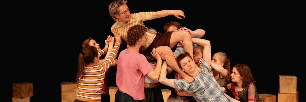 Een groep artiesten in kleurrijke casual kleding tillen en ondersteunen een persoon horizontaal boven hen in een acrobatische podiumformatie, met houten kisten op de achtergrond.

A group of performers in colorful casual clothing lift and support a person horizontally above them in an acrobatic stage formation, with wooden boxes in the background.