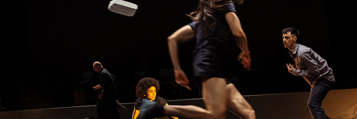 Dansers treden op op een donker podium met een live band op de achtergrond, één danser wordt opgetild terwijl anderen witte rechthoekige voorwerpen vasthouden.

Dancers perform on a dark stage with a live band in the background, one dancer being lifted while others hold white rectangular objects.