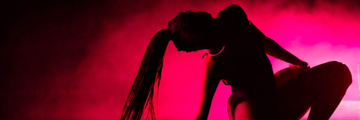 Een artiest met lange vlechten hurkt op een donker podium onder felroze verlichting, in hoge hakken en een bloot kostuum met uitwaaiend haar.

A performer with long braids crouches on a dark stage under vibrant pink lighting, wearing high heels and a revealing costume with flowing hair.