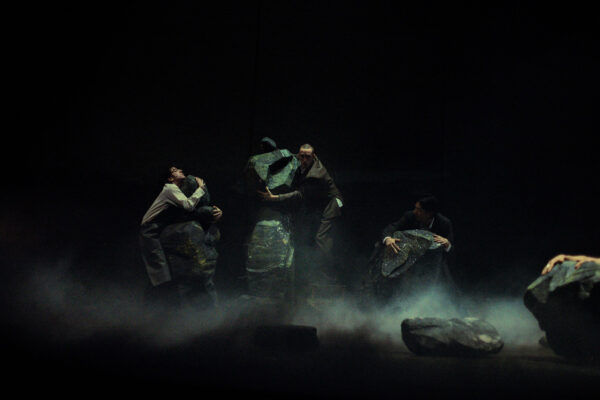 Vier performers in donkere kostuums houden grote, getextureerde stenen vast op een donker, rokerig podium, een sfeervolle theaterscène.

Four actors in dark costumes hold large, textured rocks on a dimly lit, smoky stage, creating an intense dramatic atmosphere.