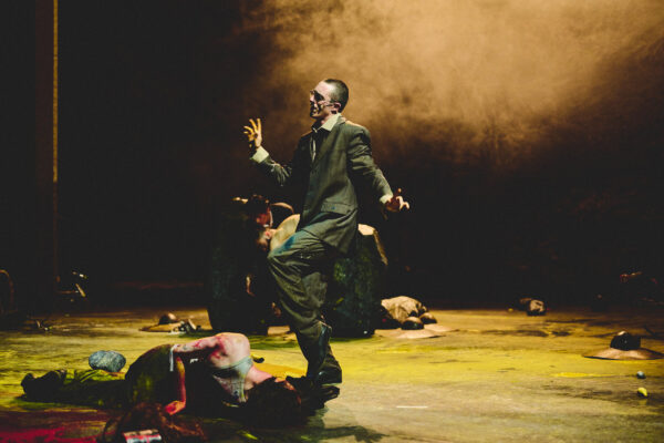 Een man in een donker pak gebaart dramatisch op een donker podium met intense, sfeervolle lichteffecten en schaduwen.

A man in a dark suit gestures dramatically on a dimly lit stage with intense, moody lighting and shadows.