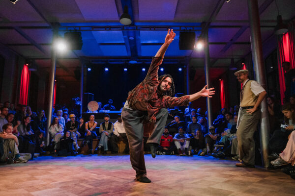 Een danser in een patronenhemd treedt op op een houten dansvloer met een aandachtig publiek dat op de achtergrond zit.

A dancer in a patterned shirt performs on a wooden stage with an attentive audience seated in the background.