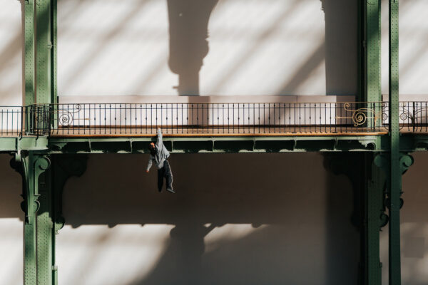 Persoon hangt aan sierlijke groene ijzeren boog in verlichte zaal met schaduwen.

Person hanging from the green iron arch in sunlit hall with shadows