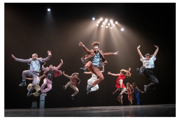 Een groep dansers in kleurrijke kostuums springt in de lucht op een donker podium onder fel licht tijdens een voorstelling.

A group of dancers in colorful costumes leaps mid-air on a dark stage under bright overhead lights during a performance.