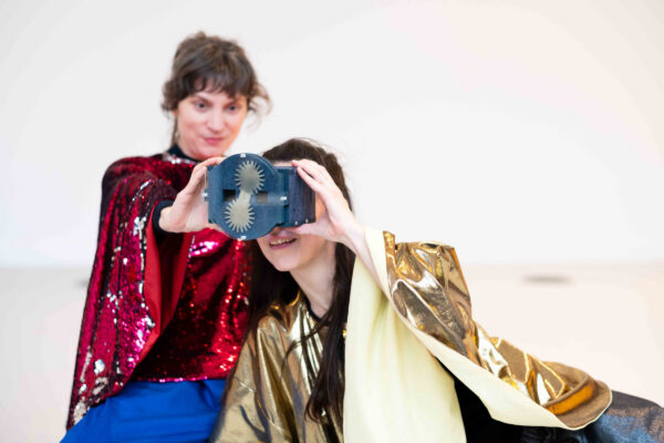 Twee personen, één in een rode paillettenjas en een andere in een gouden cape, houden een apparaat voor hun gezicht in een lichte studio.

Two people, one in a red sequin jacket and another in a gold cape, hold a device to their face in a bright studio.