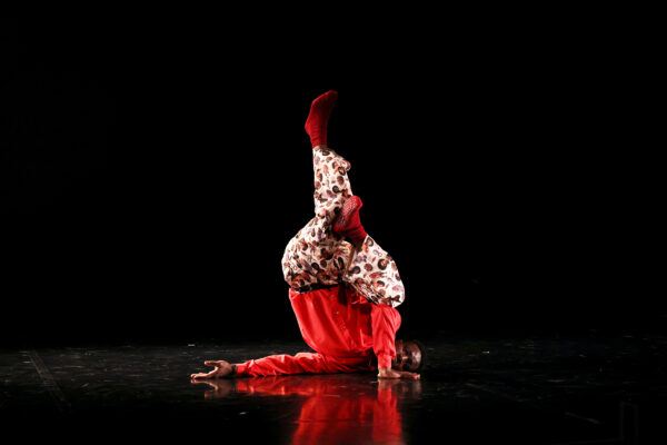 Een danser in een rood patronenpak voert een kopstand uit op een donker podium met dramatische verlichting en reflecties.

A dancer in a red patterned outfit performs a headstand on a dark stage with dramatic lighting and reflections.