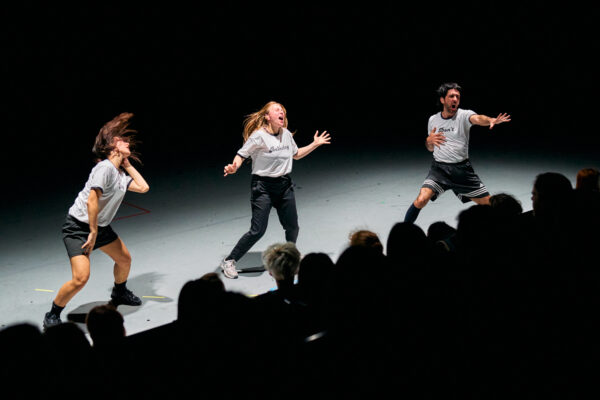 Drie dansers treden op op een donker podium met publiek op de achtergrond, twee in witte shirts en één in zwarte short.

Three dancers perform on a dark stage with an audience in the background, two in white shirts and one in black shorts.