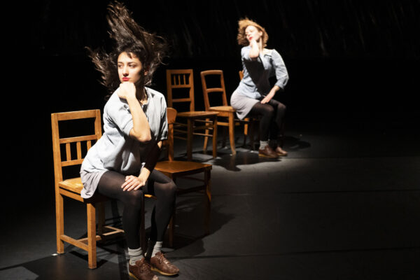 Twee vrouwen op een donker podium, één zittend op een stoel met kin in hand, de andere zit verder achterin op een stoel met wapperend haar.

Two women on a dark stage, one seated on a chair with chin in hand, the other sitting further on a chair with hair blowing.