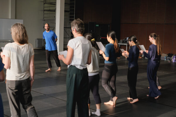 In een dansstudio observeert een instructeur in blauw shirt een rij dansers, op blote voeten of sokken, die papieren vasthouden en luisteren.

In a dance studio, an instructor in a blue shirt watches a line of dancers, barefoot or in socks, holding papers and listening attentively.