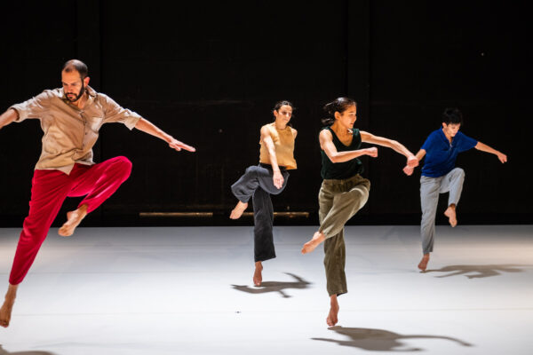 Vier dansers voeren dynamische poses uit op een donker podium met gekleurde lijnen op de vloer, gekleed in sportieve kleding.

Four dancers perform dynamic poses on a dark stage with colored floor lines, wearing casual athletic clothing.
