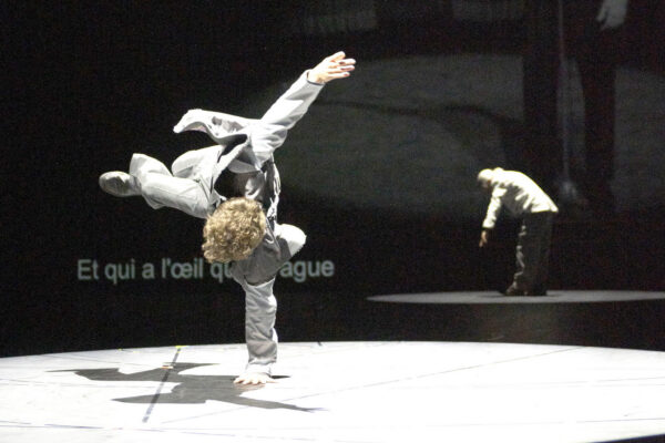 Een danser in een wit vechtkunstpak voert een hoge kick uit op een donker podium met een andere persoon en tekst op de achtergrond.

A dancer in a white martial arts outfit performs a high kick on a dark stage with another person and text in the background.