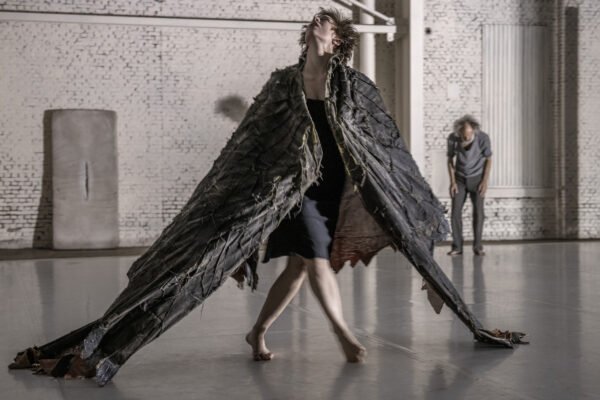 Een persoon in een versleten zwart vleugelachtig kostuum loopt in een witte bakstenen studio met een andere persoon op de achtergrond.

A person in a tattered black wing-like costume walks in a white brick-walled studio with another person in the background.