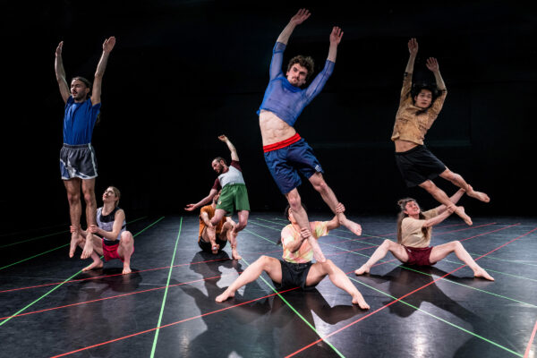 Een groep dansers voert acrobatische liften en dynamische poses uit op een donker podium met rode en groene vloerlijnen, gekleed in sportieve shorts en shirts, met opvallende houdingen.

A group of dancers performs acrobatic lifts and dynamic poses on a dark stage with red and green floor tape, wearing athletic shorts and shirts, creating striking shapes.