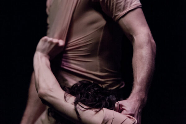 Een persoon in een roze kostuum tilt een andere danser ondersteboven op een donker podium met dramatische verlichting, schaduwen en reflecties.

A person in a pinkish costume lifts another dancer upside down on a dark stage with dramatic lighting, shadows, and reflections.