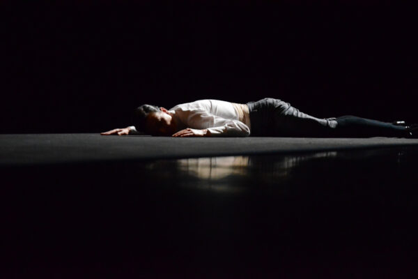 Een persoon ligt op de buik op een donker podium met uitgestrekte armen, gekleed in een wit shirt en donkere broek, onder subtiele, gerichte verlichting.

A person lies on their belly on a dark stage with arms stretched out, wearing a white shirt and dark pants, under subtle, focused lighting.