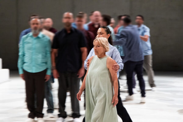 Een groep mensen in casual kleding loopt op een lichtkleurig podium, met een vrouw in een lichte groene jurk duidelijk op de voorgrond.

A group of people in casual clothing walks on a light-colored stage, with a woman in a light green dress prominently in the foreground.