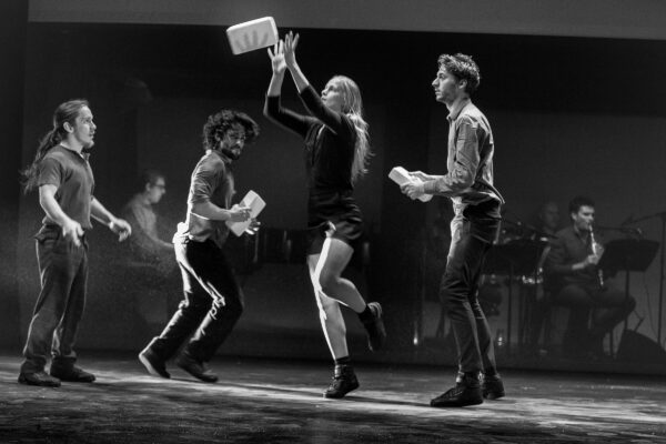 Dansers treden op op een donker podium met een live band op de achtergrond, één danser wordt opgetild terwijl anderen witte rechthoekige voorwerpen vasthouden.

Dancers perform on a dark stage with a live band in the background, one dancer being lifted while others hold white rectangular objects.