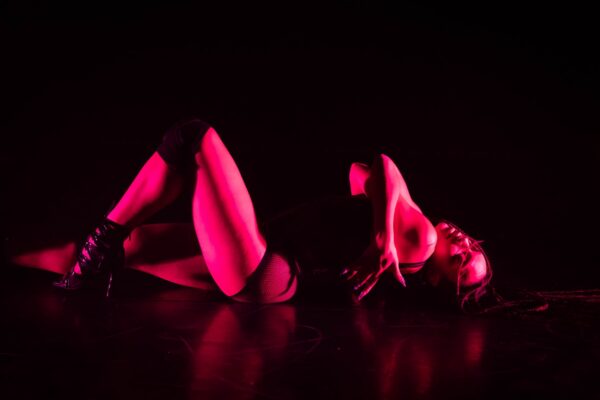 Een performer in zwarte kleding met visnetkousen ligt op de rug, benen omhoog gestrekt, badend in felroze podiumlicht.

A performer in black outfit with fishnet stockings lies on their back, legs raised high, bathed in vibrant pink stage lighting.