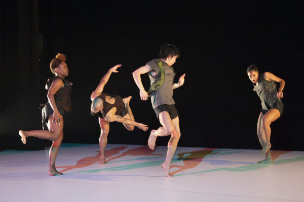 Vier dansers in donkere sportkleding voeren dynamische, atletische poses uit op een podium met kleurrijke abstracte vloerprojecties in rood, blauw en groen.

Four dancers in dark athletic wear perform dynamic, athletic poses on a stage with colorful abstract floor projections in red, blue, and green.
