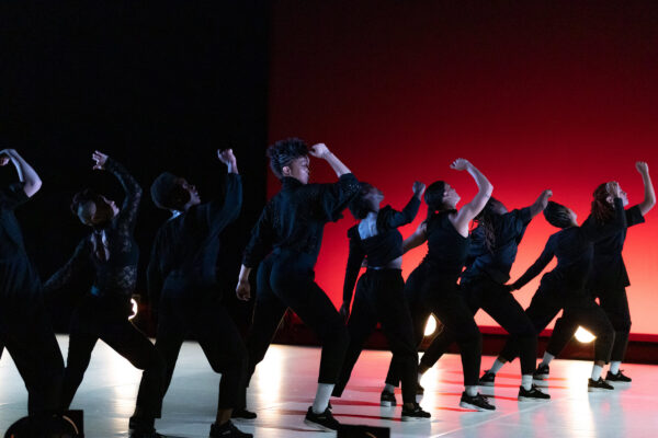 Een groep dansers in zwarte kostuums treden op met opgeheven armen op een podium met een rode gradient achtergrond, dramatische verlichting en schaduwen.

A group of dancers in black costumes perform with arms raised on a stage with a red gradient backdrop, dramatic lighting, and shadows.