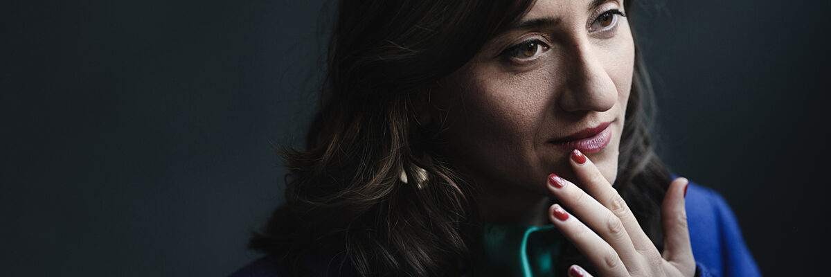 Portret van een vrouw met lang donker golvend haar in een blauw blazer, hand bij haar kin, tegen een donkere wazige achtergrond.