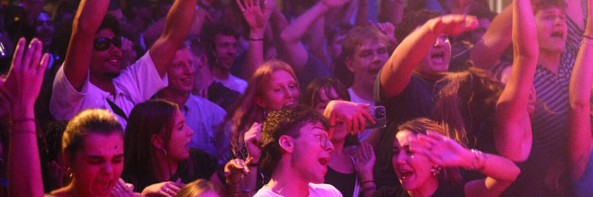 Een energieke menigte jonge volwassenen die enthousiast samen dansen op een feest met fel paarse en roze podiumverlichting, handen omhoog in vreugdevolle viering

English: An energetic crowd of young adults dancing enthusiastically together at a party with vibrant purple and pink stage lighting, hands raised in joyful celebration