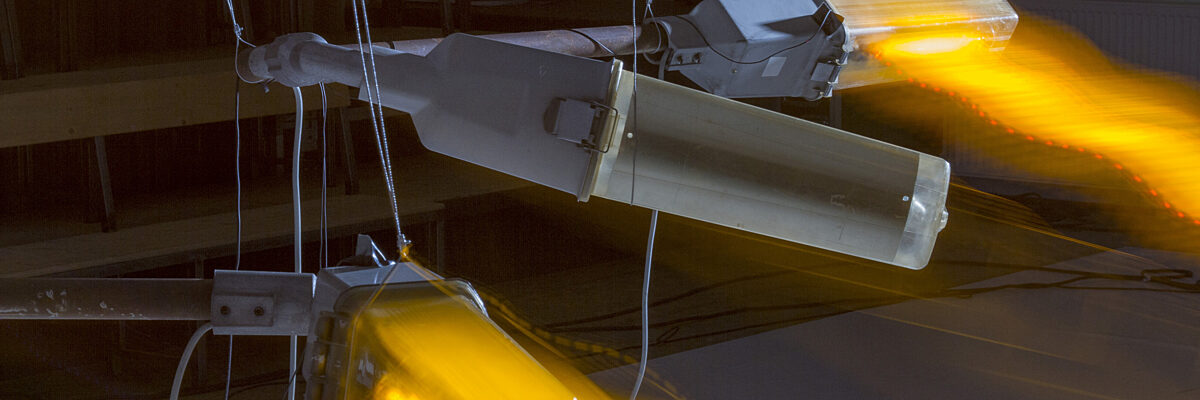 Industrial fixture with bright orange light, suspended by cables in a dark room.