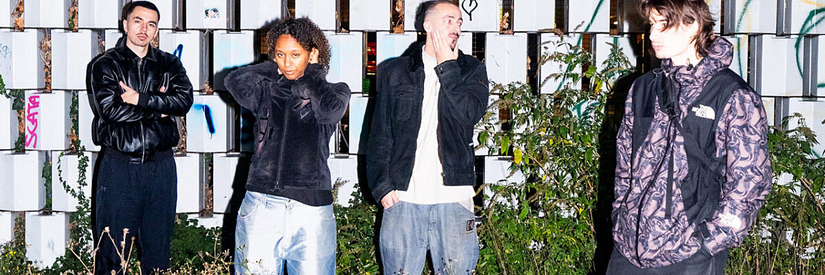 Four people stand in front of a patterned white-and-gray block wall, surrounded by overgrown grass.