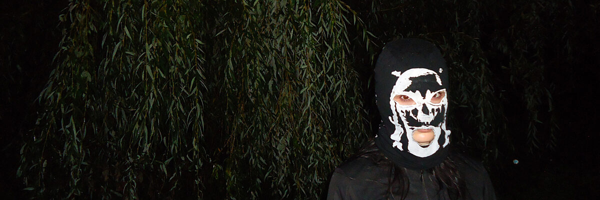 Persoon draagt een zwart masker met een witte schedelprint, staand voor donkere bomen met hangende takken.