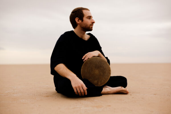 Person sits on the ground in a sandy environment, holding a large hand drum and gazing into the distance.